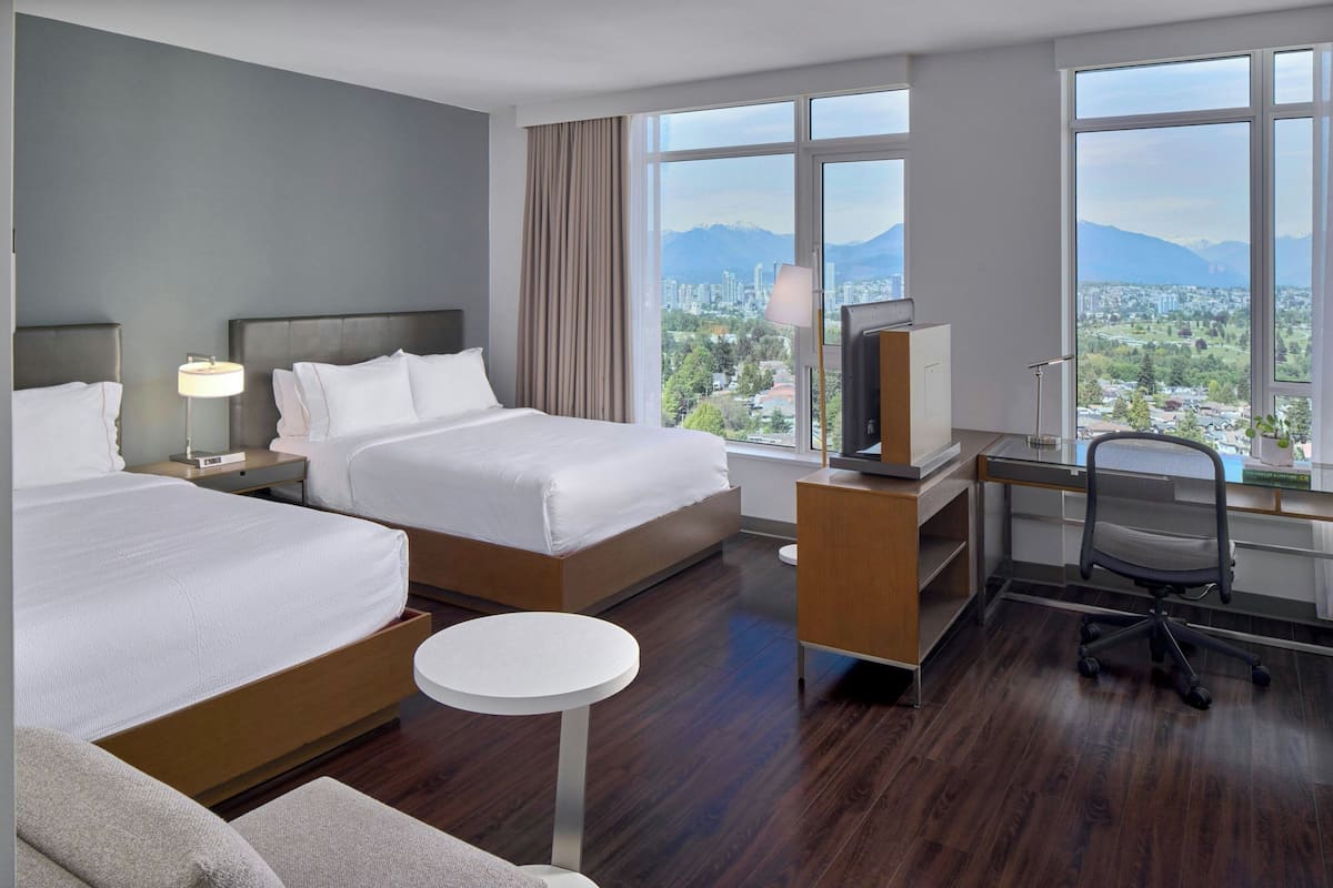 A hotel room with two beds, a desk, a chair, a small round table, and a TV.