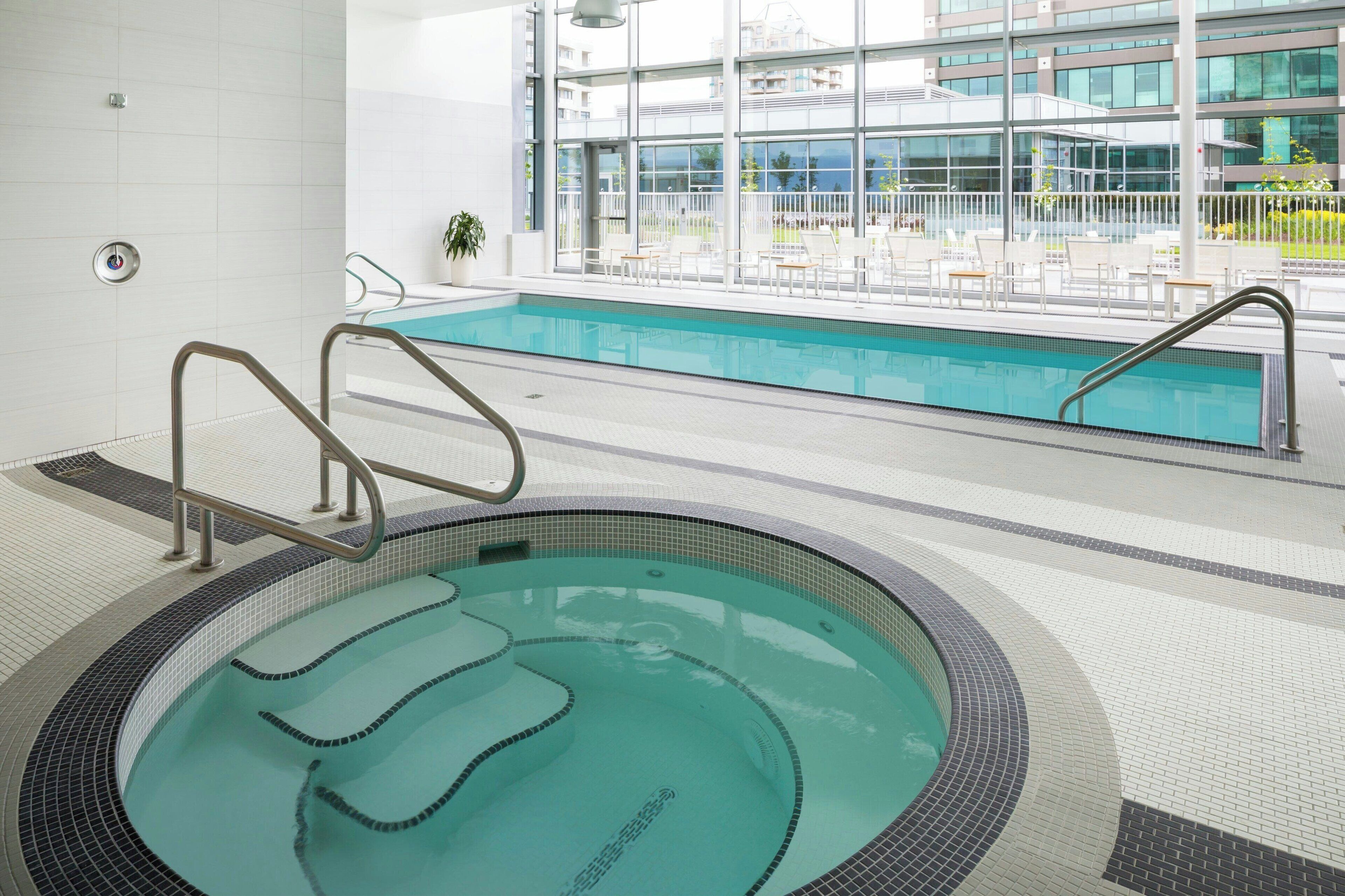 A swimming pool with a tiled deck, a lounge area with chairs and tables, and a view of a modern building with large windows.