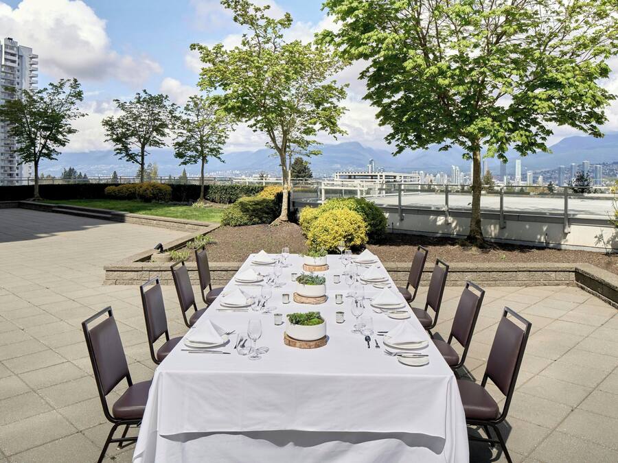 An outdoor dining area with a long table set for a meal, surrounded by chairs, with a cityscape and mountains in the background.