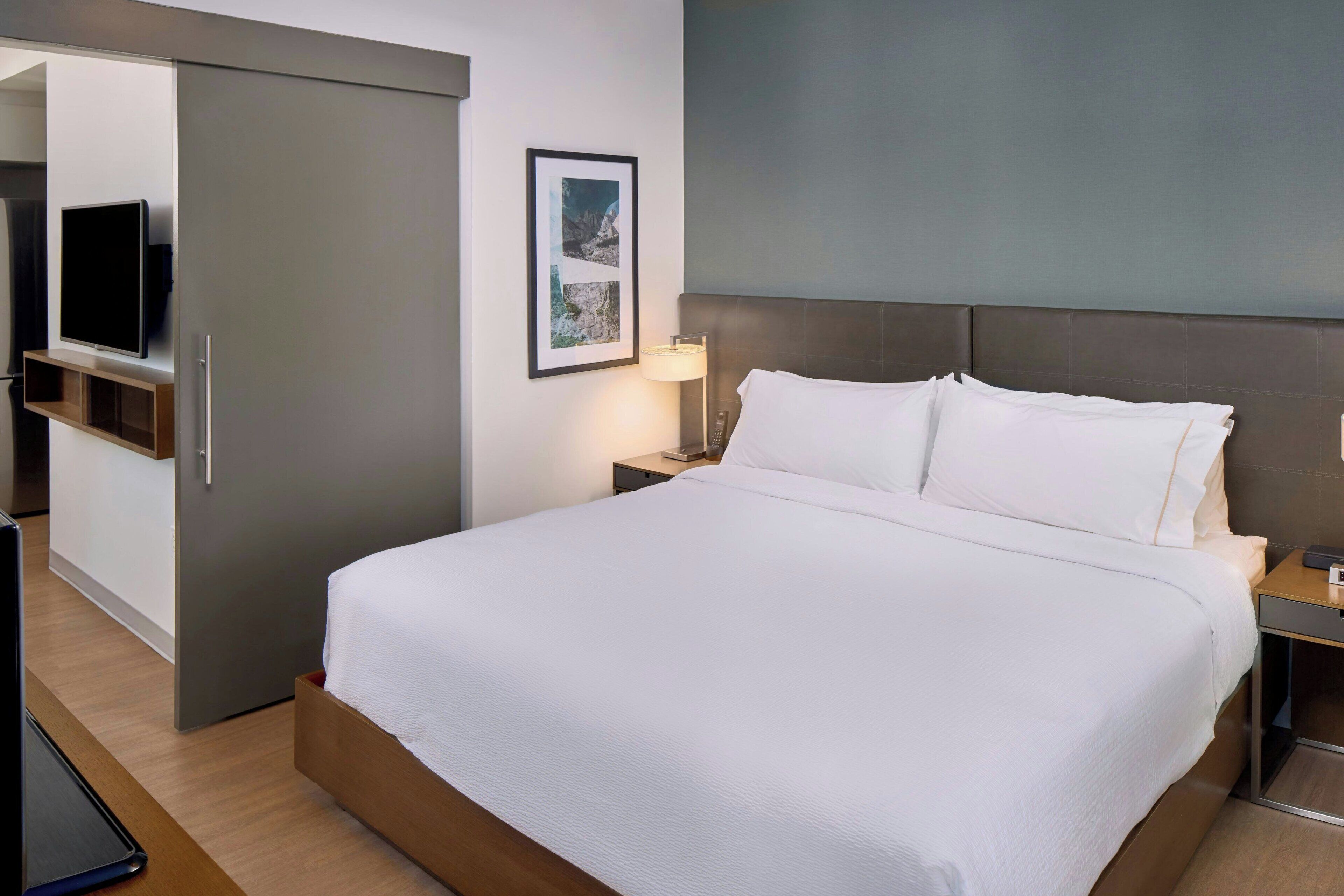 Une chambre d’hôtel avec un grand lit, un téléviseur posé sur un meuble, une table de chevet avec une lampe et un tableau encadré au mur.