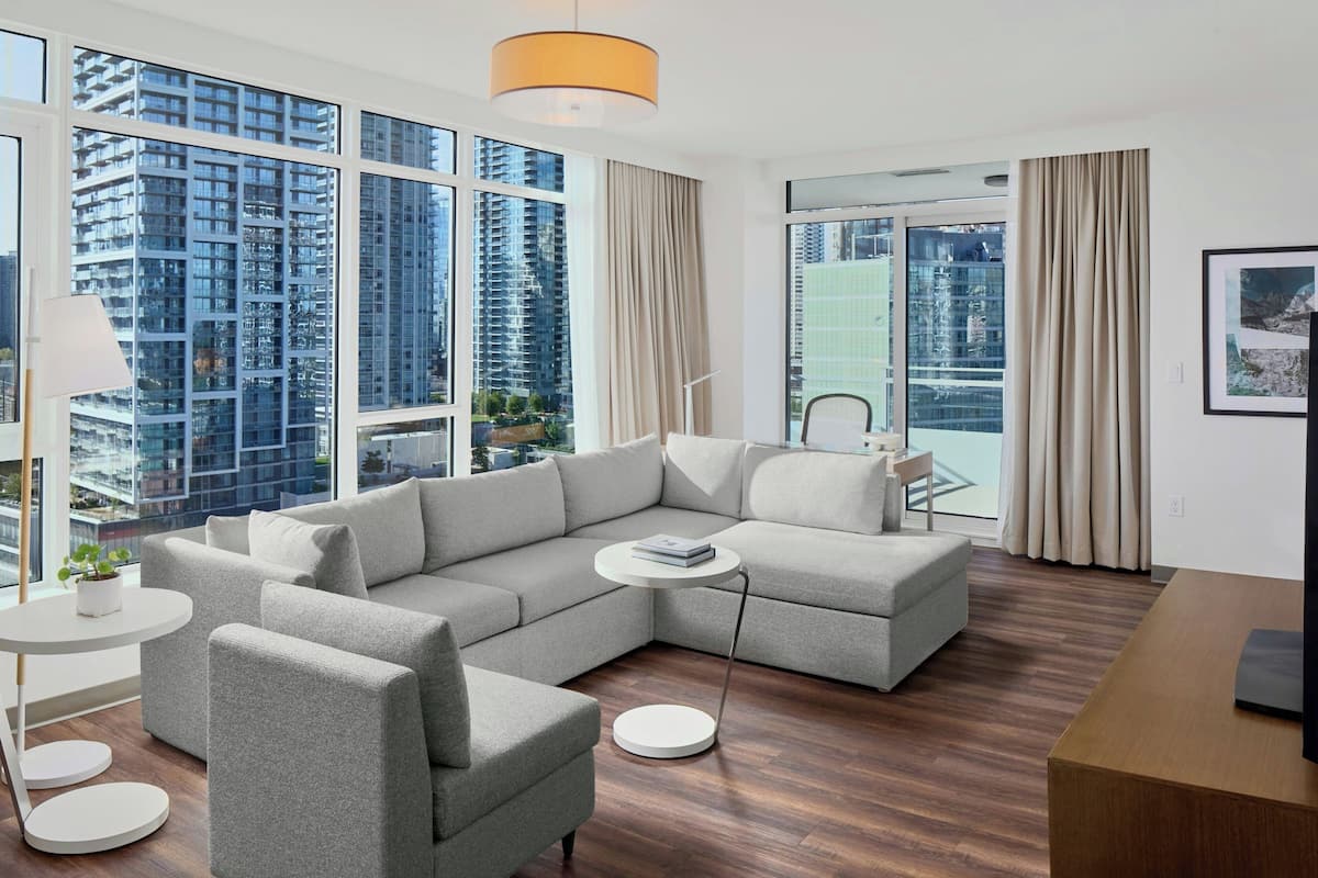 A modern living room with a sectional sofa, a round coffee table, and a TV stand. Large windows offer a view of high-rise buildings.