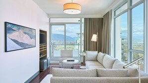 Una sala de estar moderna con un sofá, una mesa de centro y una televisión. Hay un ventanal con vistas a la ciudad y las montañas.