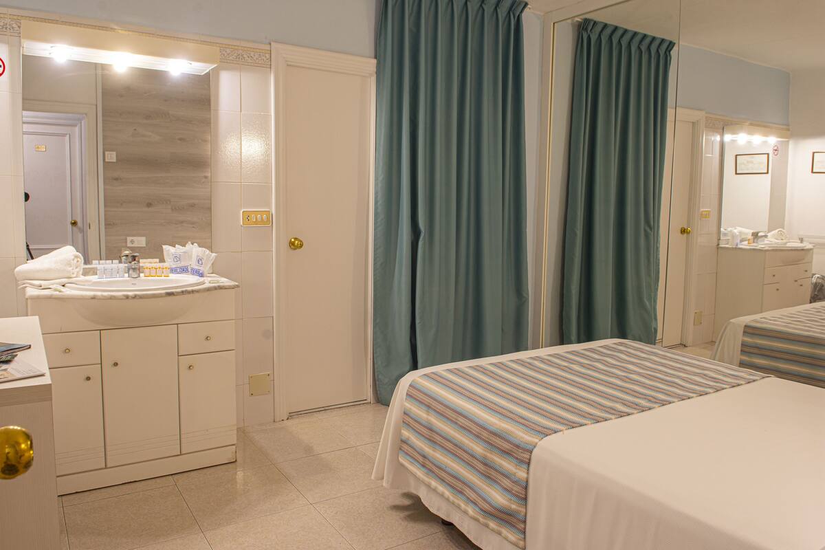 A hotel room with a bed, a bedside table, a mirror, a sink, and curtains.