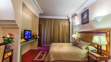 Habitación de hotel con una cama grande, un escritorio con televisor, una mesita de noche con lámpara y alfombra roja.