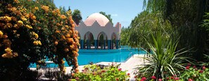 Un espace piscine agrémenté d’un bâtiment doté d’un dôme et d’arcades, le tout entouré d’une végétation luxuriante et de plantes en fleurs.