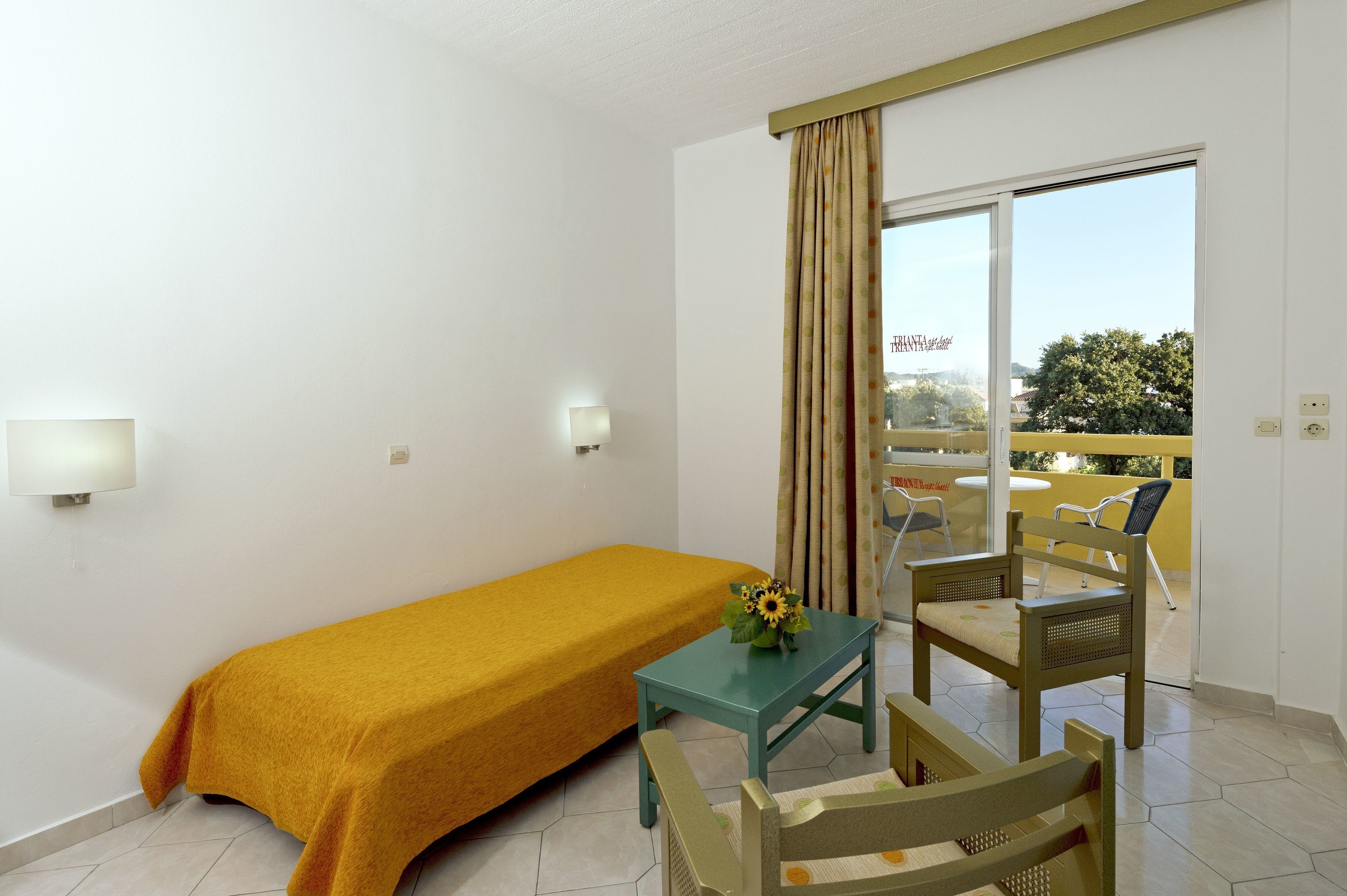 A hotel room with a single bed, a small table with a flower, a chair, and a balcony with a yellow railing.