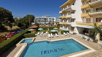 A swimming pool with 'TRIANTA' written on the water, surrounded by lounge chairs and umbrellas, with a building in the background.