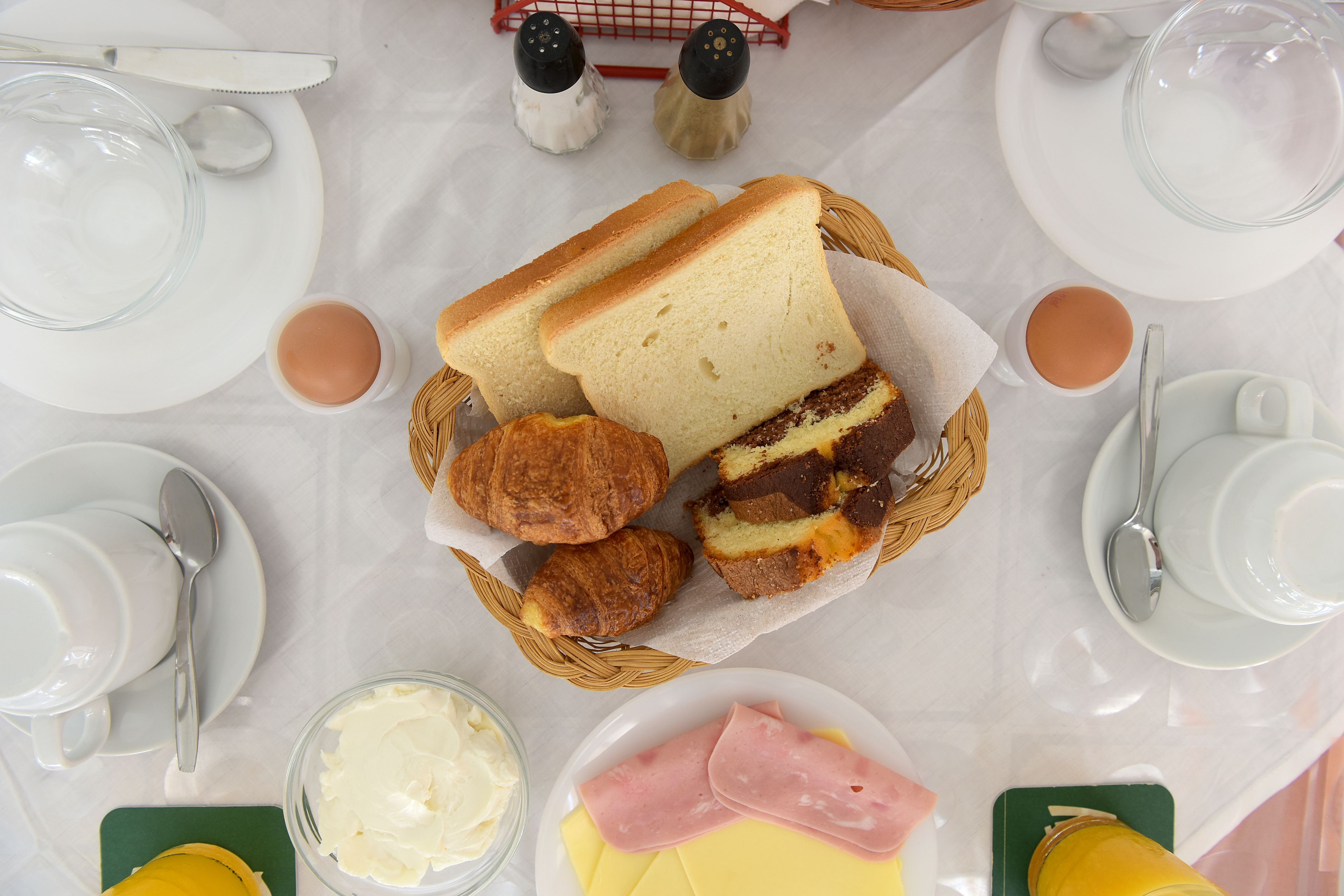 Mesa posta para o café da manhã com pão, doces, manteiga, queijo, presunto e um copo de suco de laranja.