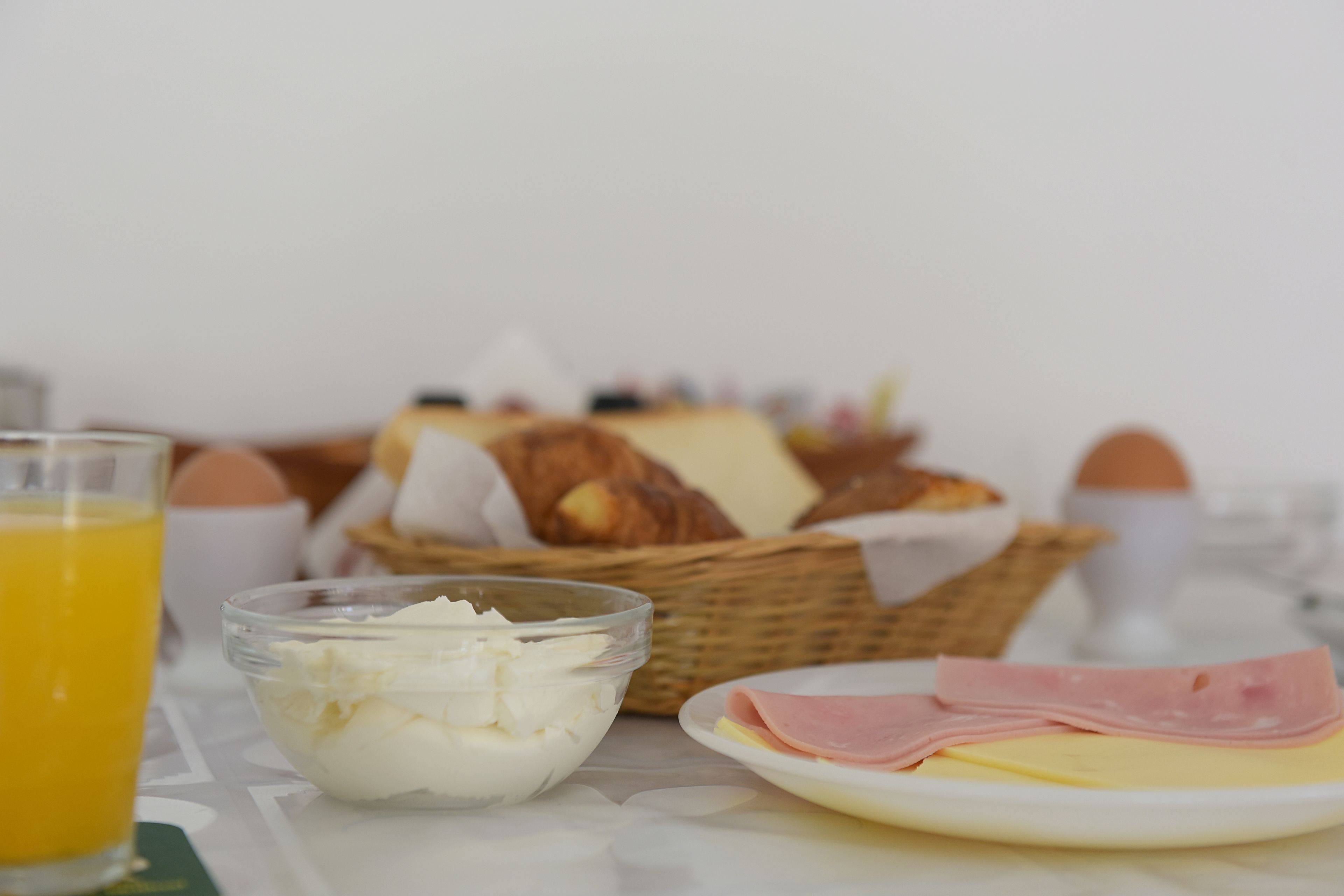 Una mesa de desayuno con un vaso de jugo de naranja, un bol de queso crema, un plato con jamón y queso, una cesta de pan y una cesta de huevos.