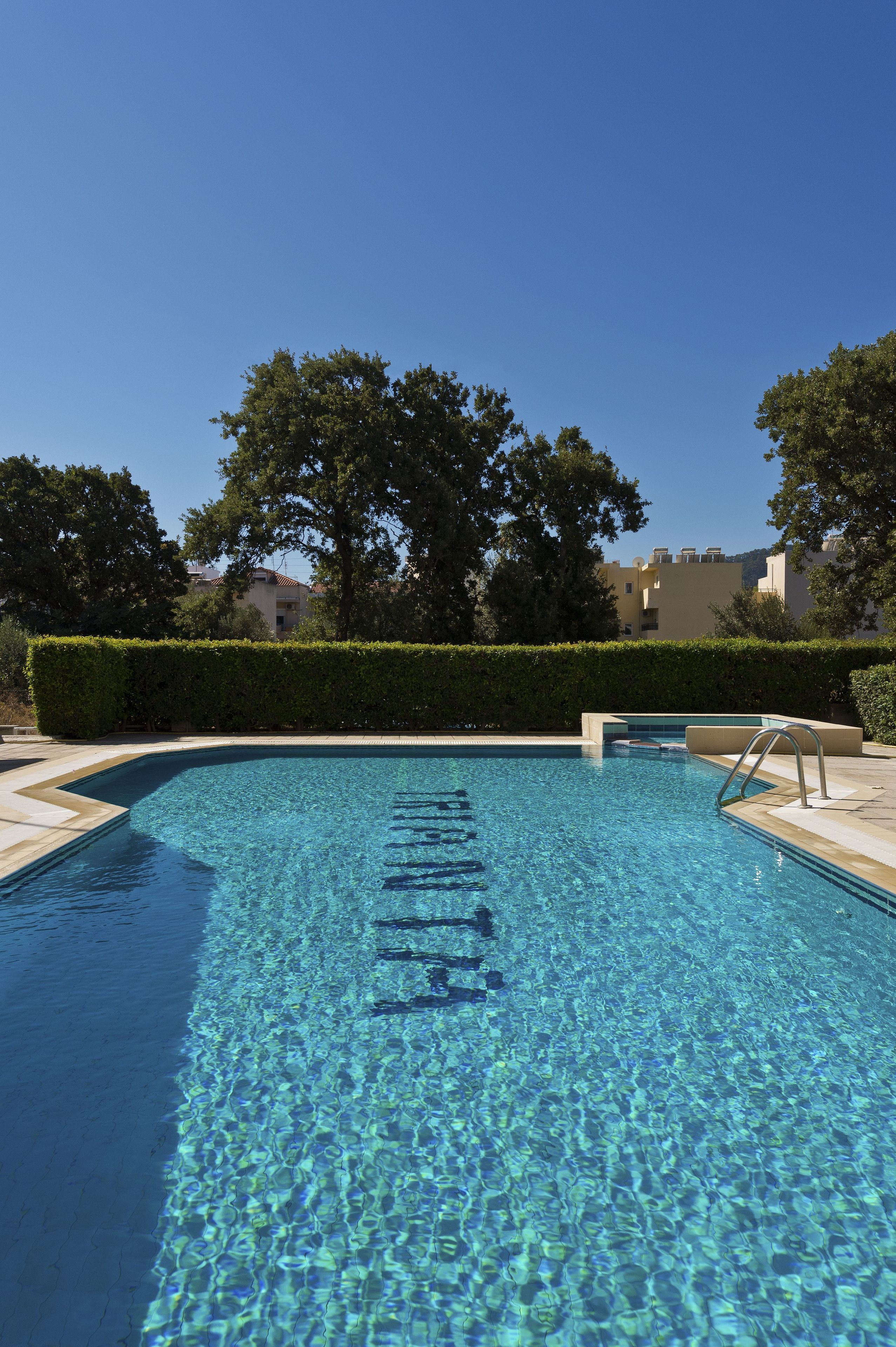 Una piscina cristalina con una escalera de metal, rodeada de setos y árboles.