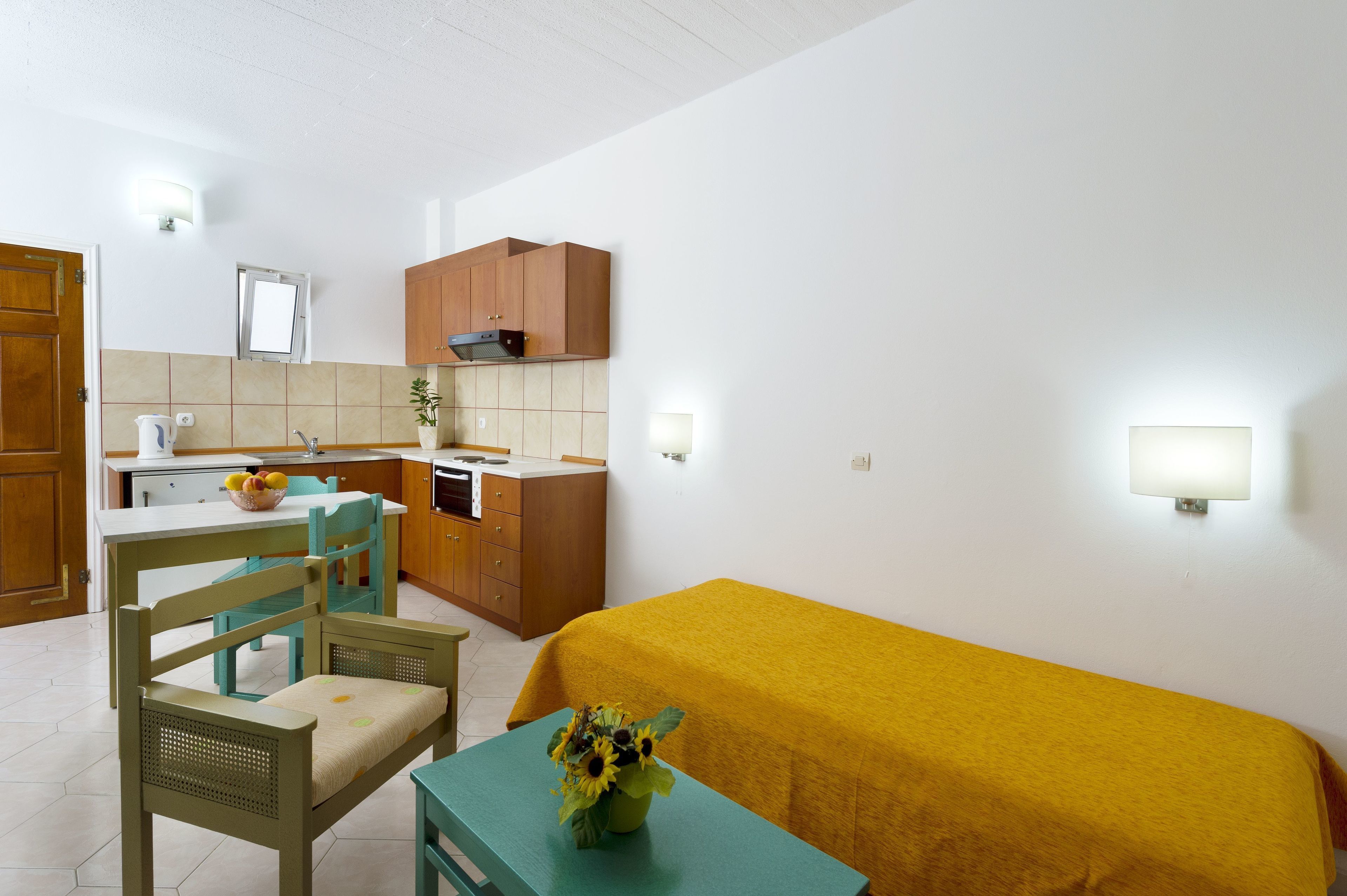 Une cuisine compacte avec des meubles en bois, un évier et une cuisinière. On y trouve un coin repas avec une table et des chaises, ainsi qu’un lit recouvert d’une couverture jaune.