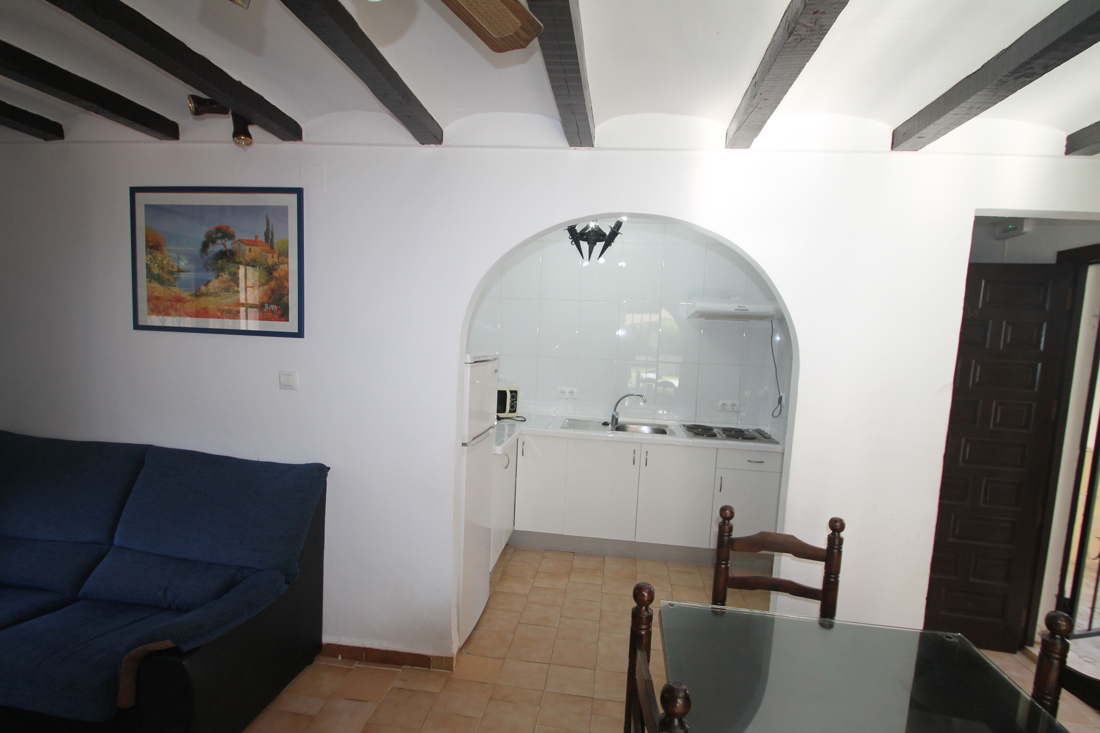 Una sala de estar con un sofá azul, una mesa de comedor con sillas, una cocina con armarios blancos y un piso de baldosas.