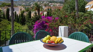 Une table avec un saladier de fruits et une nappe blanche, entourée de chaises vertes, avec vue sur un paysage montagneux et des bâtiments en arrière-plan.