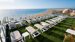 A rooftop terrace with rows of white lounge chairs and tables, overlooking a beach and ocean. - Los Nardos (San Bartolome de Tirajana)