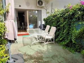 An outdoor patio with a glass table and two chairs, a covered seating area, and a green hedge wall. - Rinconada Real Bungalows (Benidorm)