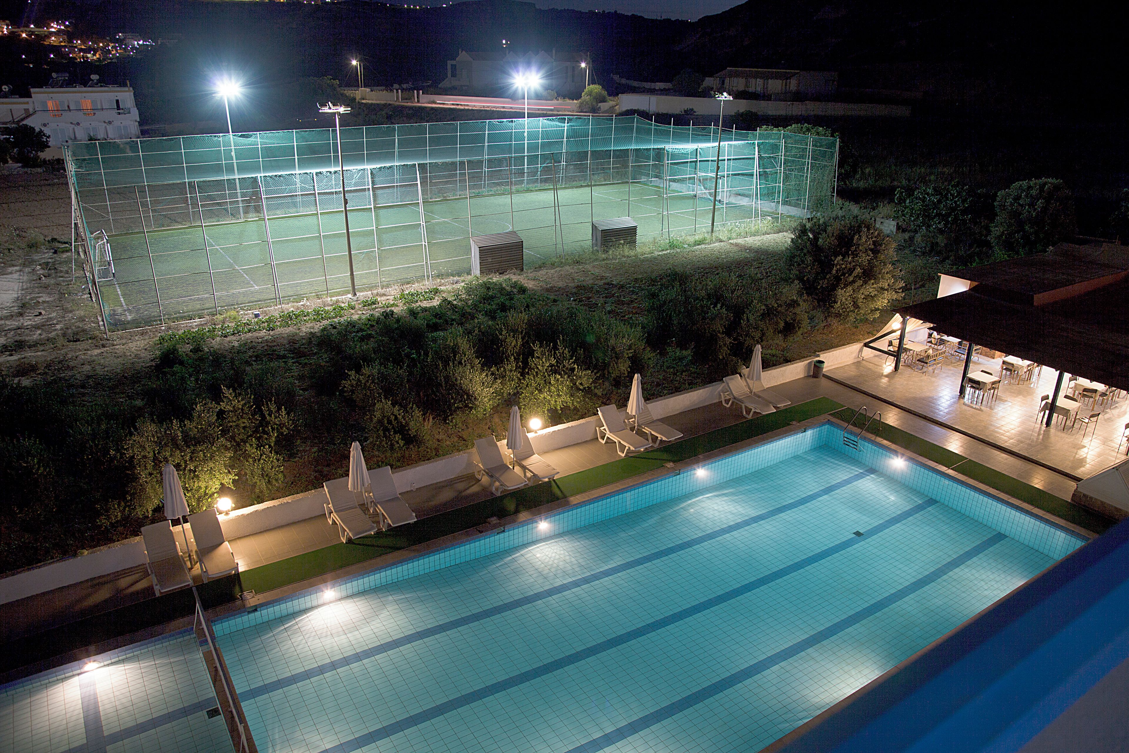 En swimmingpool med tillhörande solstolar och parasoller, omgiven av ett inhägnat område med en tennisbana.