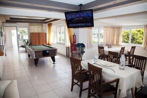 A room with a pool table, dining area with chairs, a television mounted on the wall, and a view of the outdoors through the windows. - Athinoula (Kos)