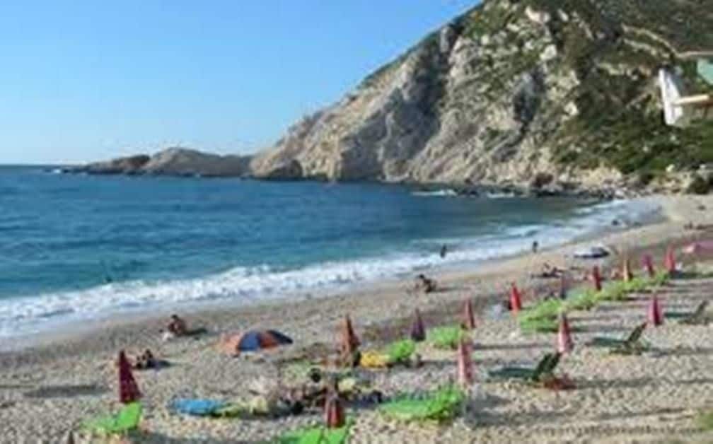 A beach with people sunbathing and swimming, a rocky cliff in the background, and umbrellas lined up along the shore.