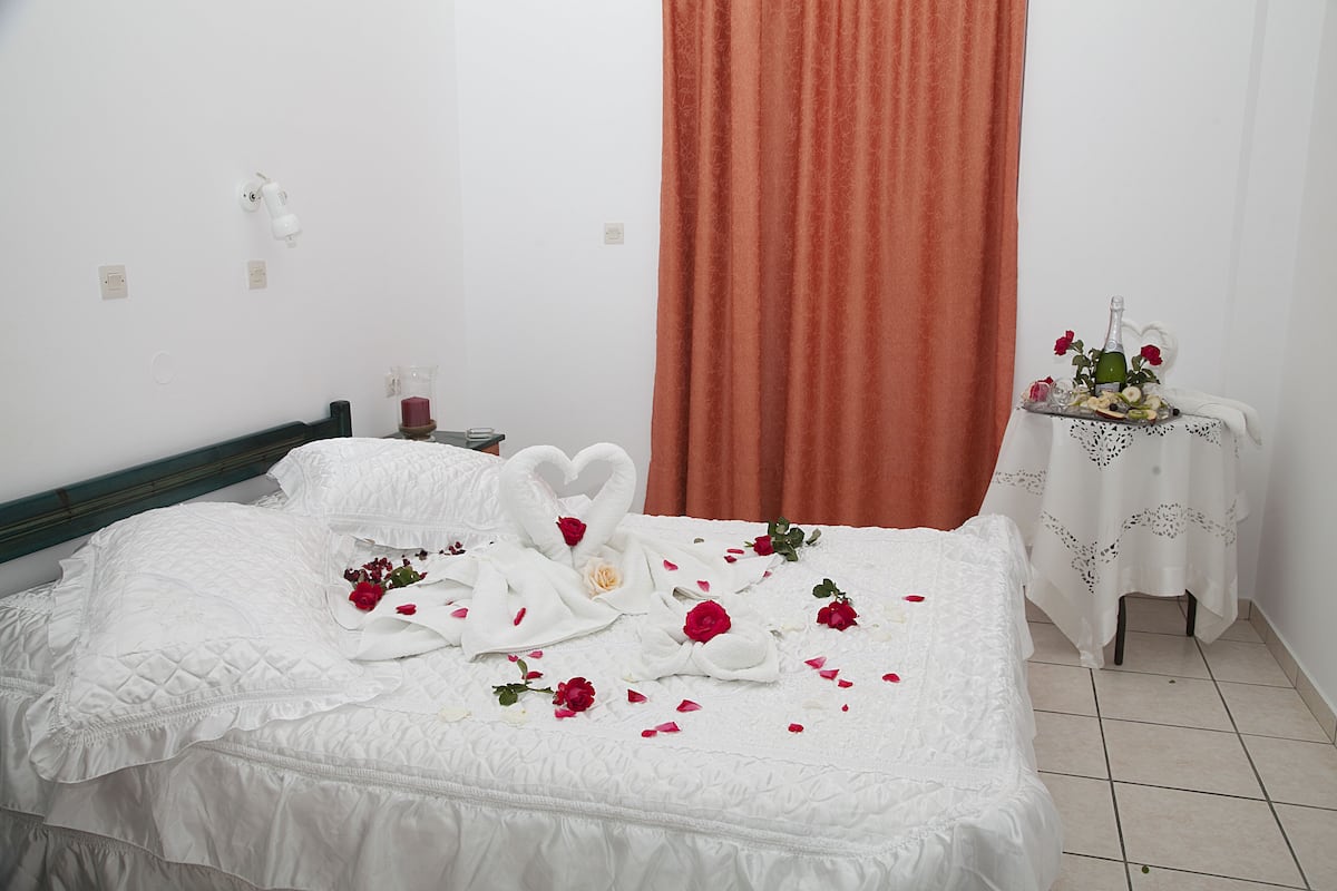 A neatly made bed with a heart-shaped towel arrangement and rose petals, a small table with a bottle and flowers, and a red curtain.