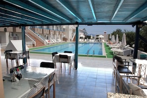 Ett utomhusområde med en pool som är delvis täckt, utrustat med solstolar och bord, en trappa som leder upp till de övre våningarna och en utsikt över en byggnad i bakgrunden.