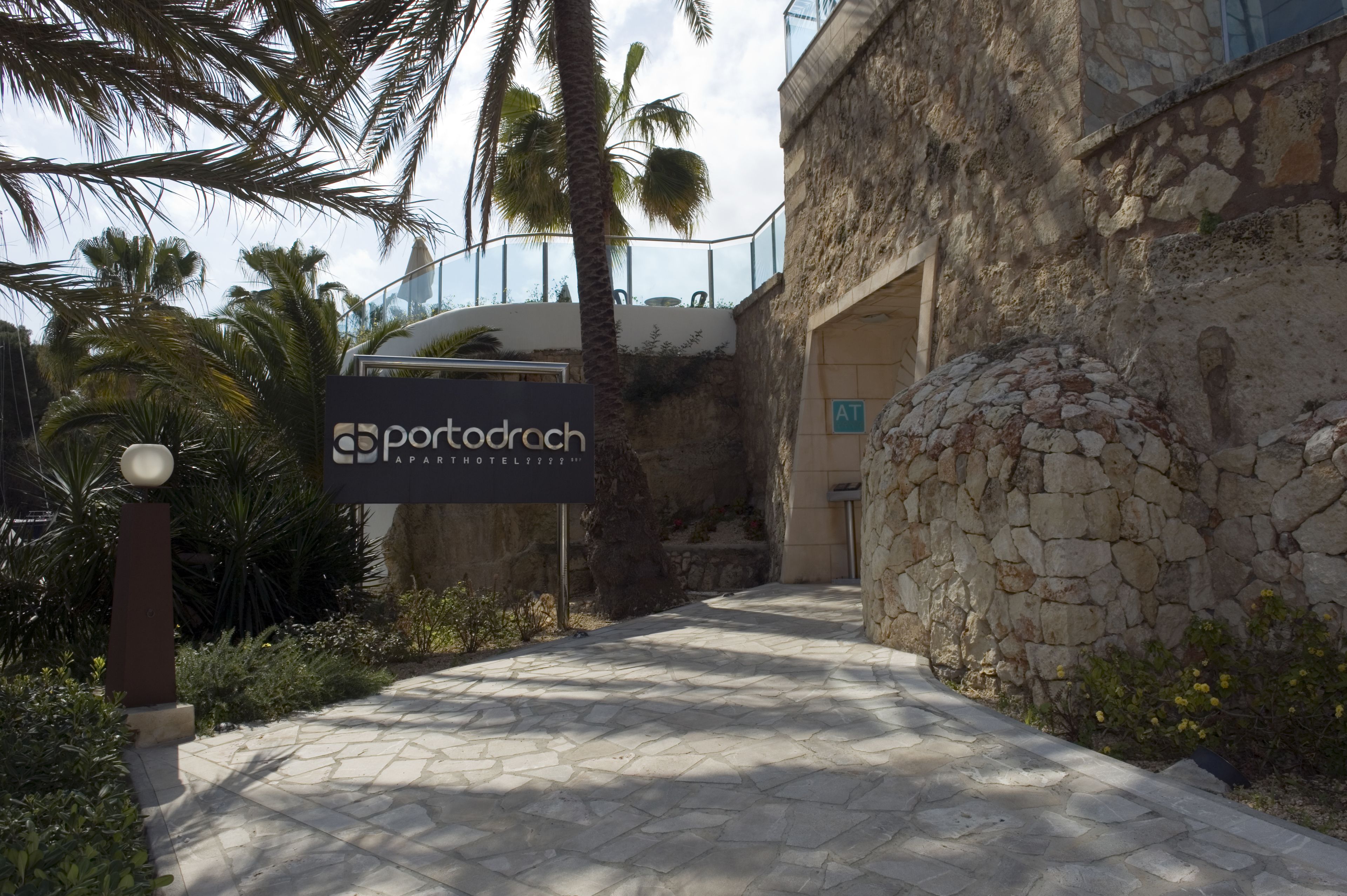 A pathway leading to a building with a stone facade and palm trees. A signboard for Portodroch Hotel is visible.