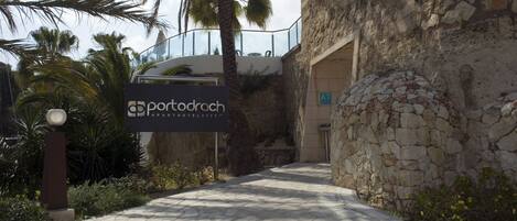 A pathway leading to a building with a stone facade and palm trees. A signboard for Portodroch Hotel is visible.