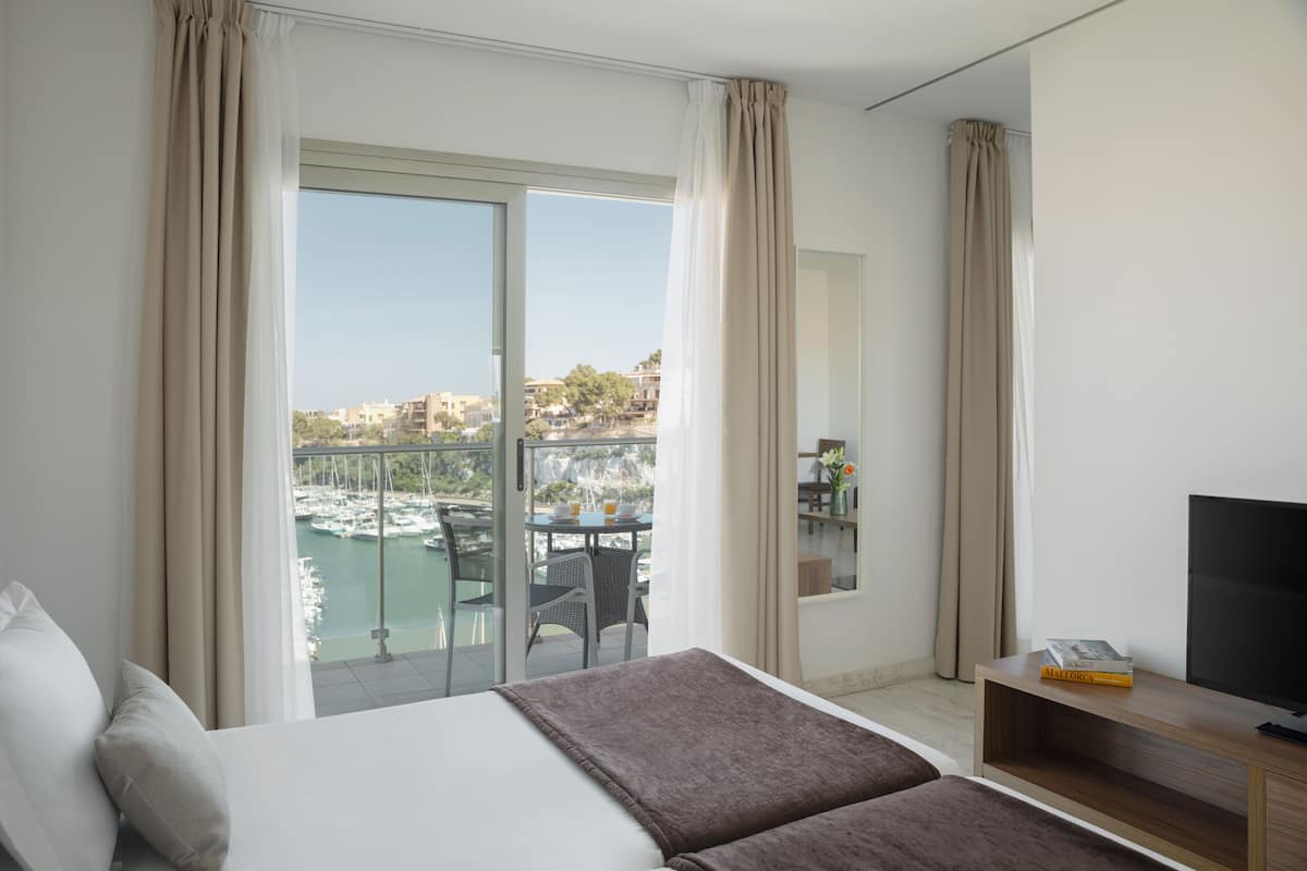 Quarto de hotel com cama, televisão, varanda com vista para a marina e área de refeições com mesa e cadeiras.