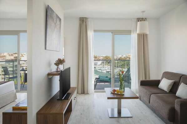 A modern hotel room with a large window, a flat-screen TV, a sofa, a coffee table, and a view of a marina. - Portodrach Aparthotel & Suites (Manacor)