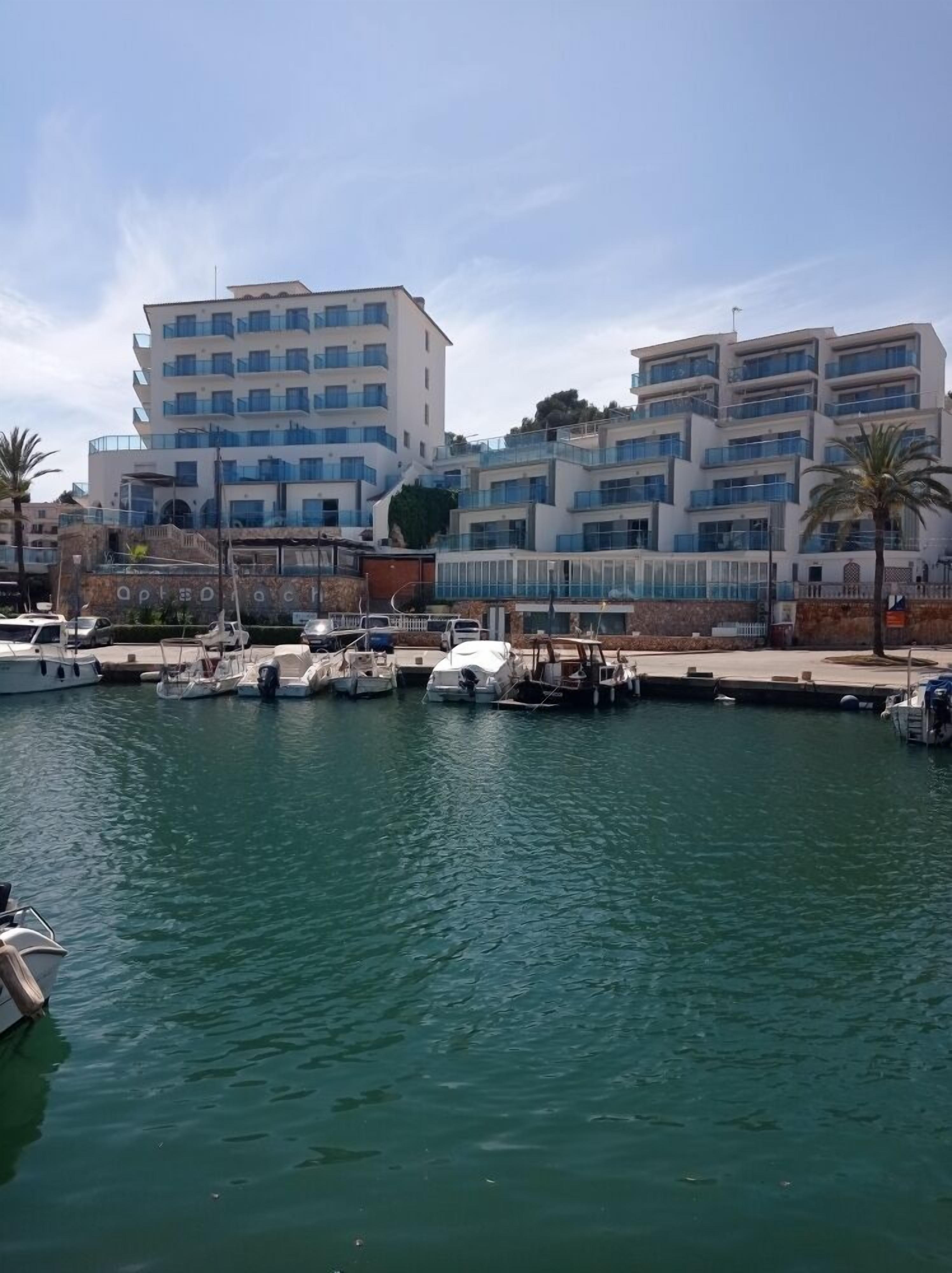 A marina with several boats docked, a modern building with multiple balconies, and a clear sky.