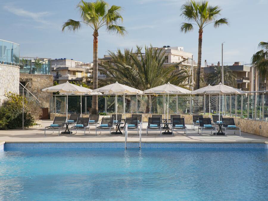 Piscina con sillas de descanso y sombrillas, rodeada de palmeras y edificios.