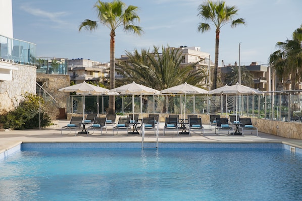 A swimming pool with lounge chairs and umbrellas, surrounded by palm trees and buildings. - Portodrach Aparthotel & Suites (Manacor)