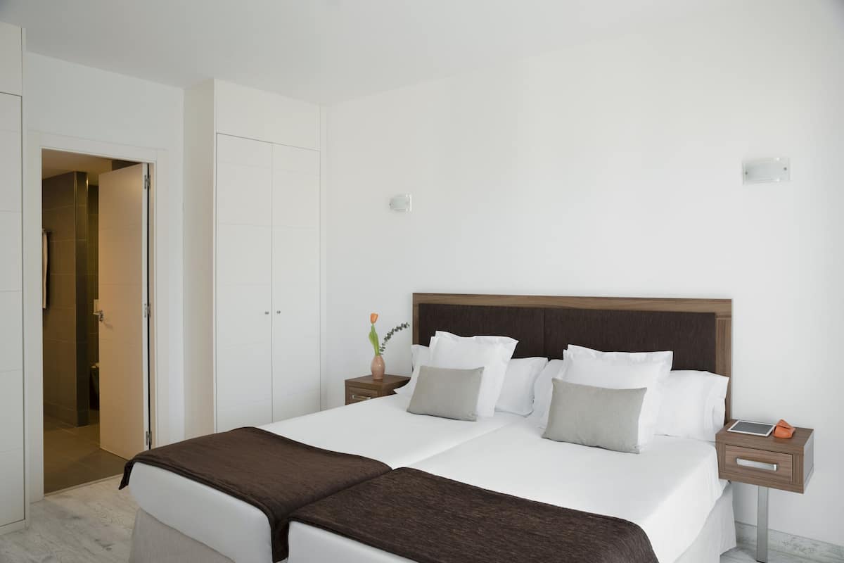 Quarto de hotel com cama grande, mesa de cabeceira, criado-mudo e guarda-roupa.