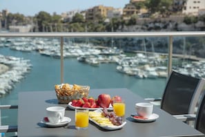A table with breakfast items, a basket of bread, and a view of a marina with boats. - Portodrach Aparthotel & Suites (Manacor)