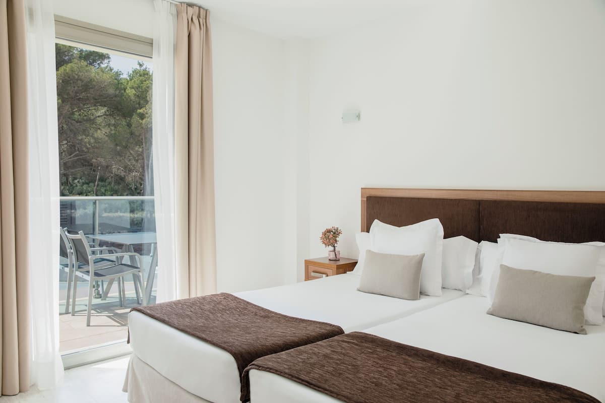 Quarto de hotel com duas camas, cabeceira de madeira, mesinha de cabeceira com um vaso e varanda com cadeiras e mesa.