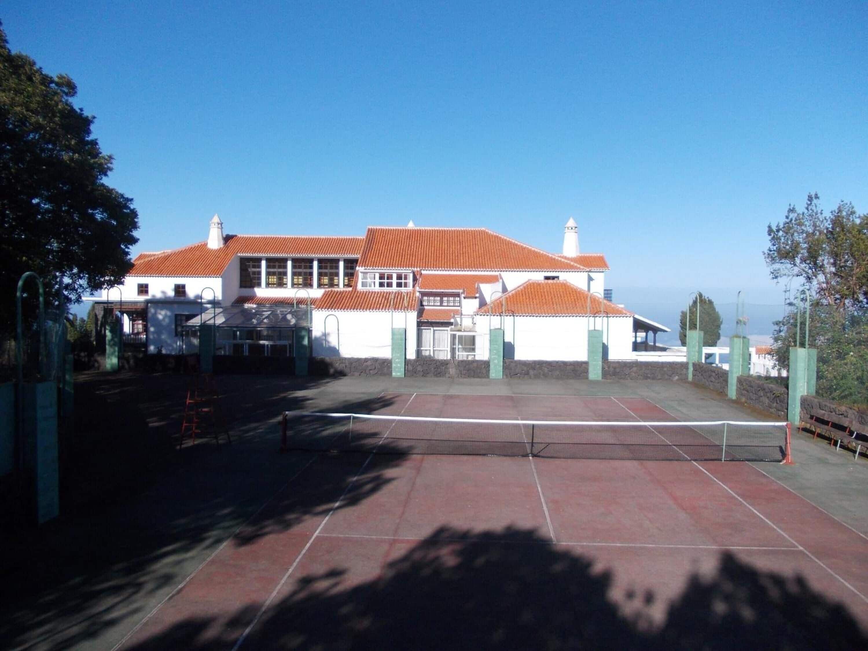 A tennis court with a net in the center, surrounded by a building with a red-tiled roof and white walls.