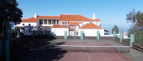 A tennis court with a net in the center, surrounded by a building with a red-tiled roof and white walls.