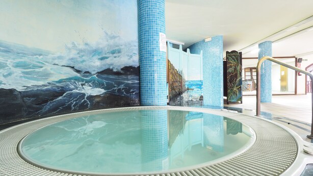 Ein Whirlpoolbereich mit einer Wandmalerei von Meereswellen, mit Fliesen versehenen Säulen und einer Glaskonstruktion, die eine Badewanne umschließt.