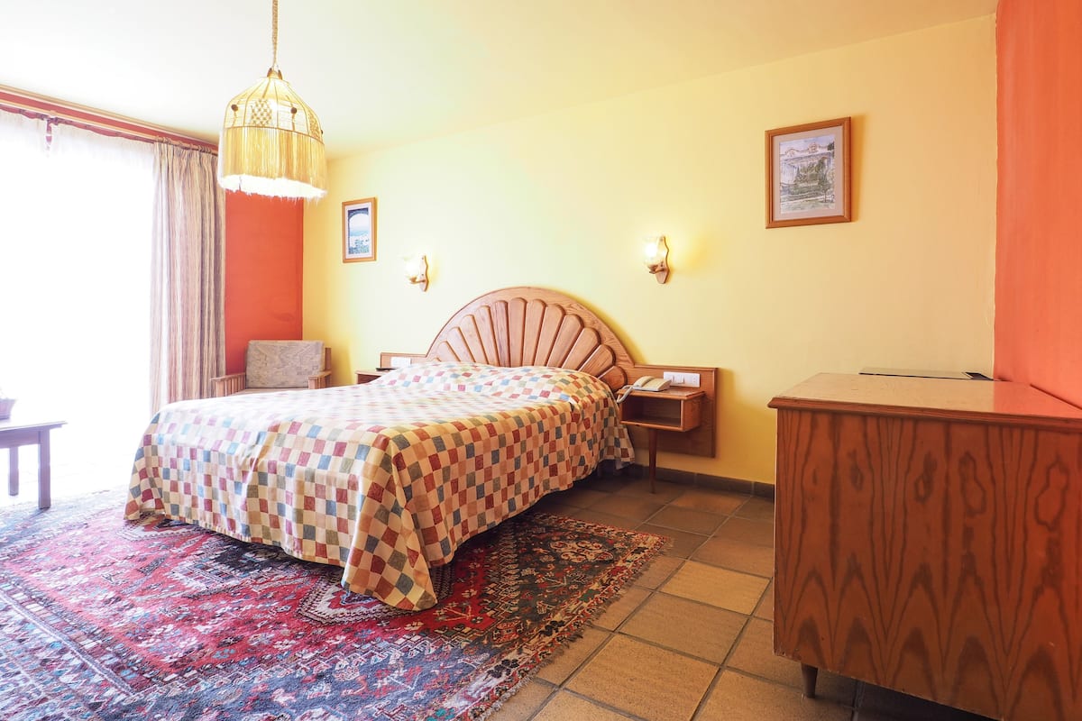 Un dormitorio con cama, mesita de noche, lámpara, cómoda de madera y alfombra estampada.