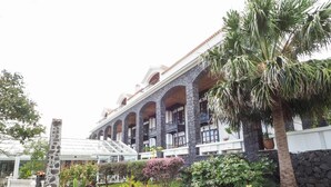 Ein Hotel mit einer Steinfassade, mehreren Balkonen und einem Garten mit verschiedenen Pflanzen und einer Palme.