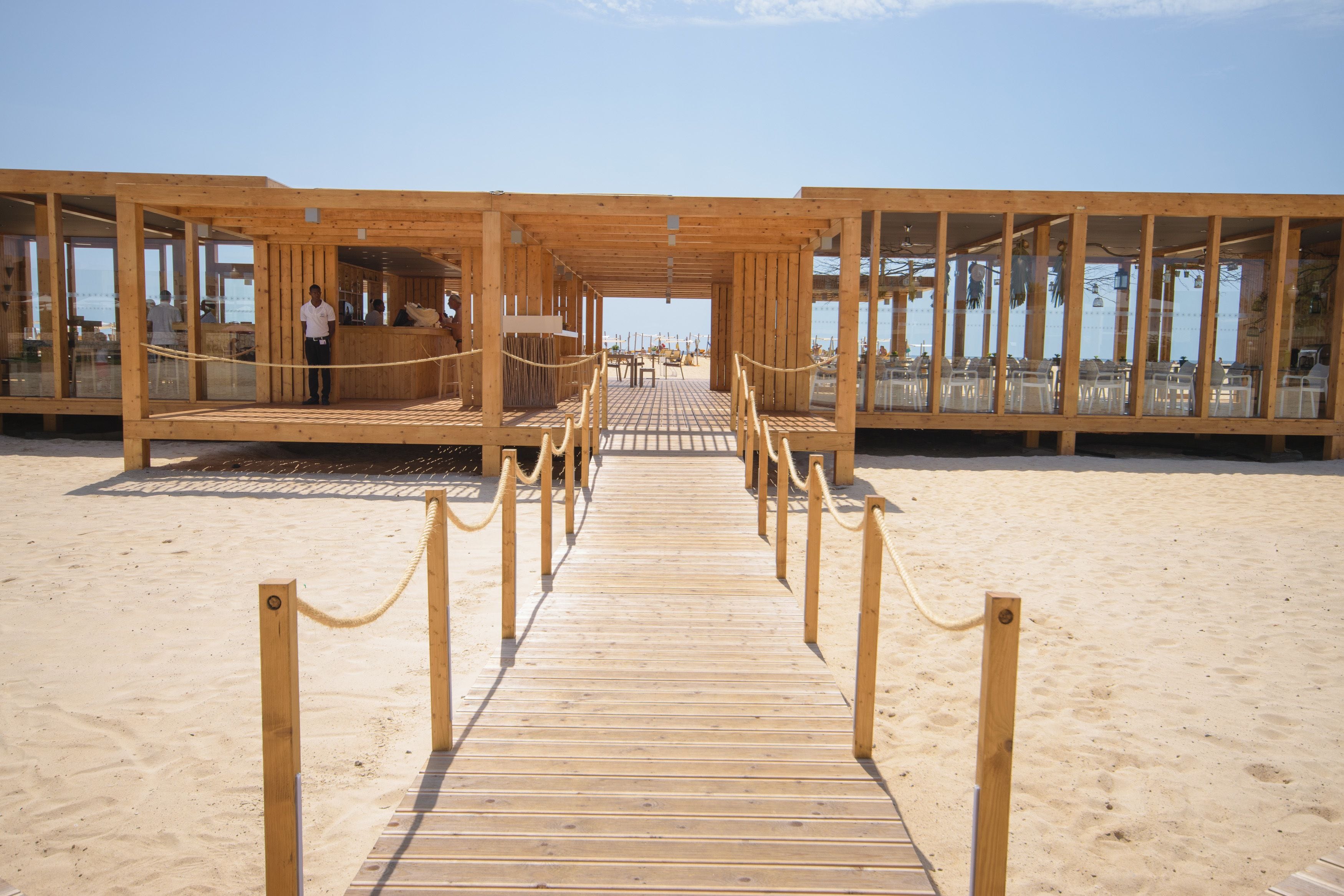 Une terrasse en bois mène à un bâtiment situé en bord de mer, doté de grandes fenêtres, d’un sol à carreaux et d’une barrière faite de cordes.