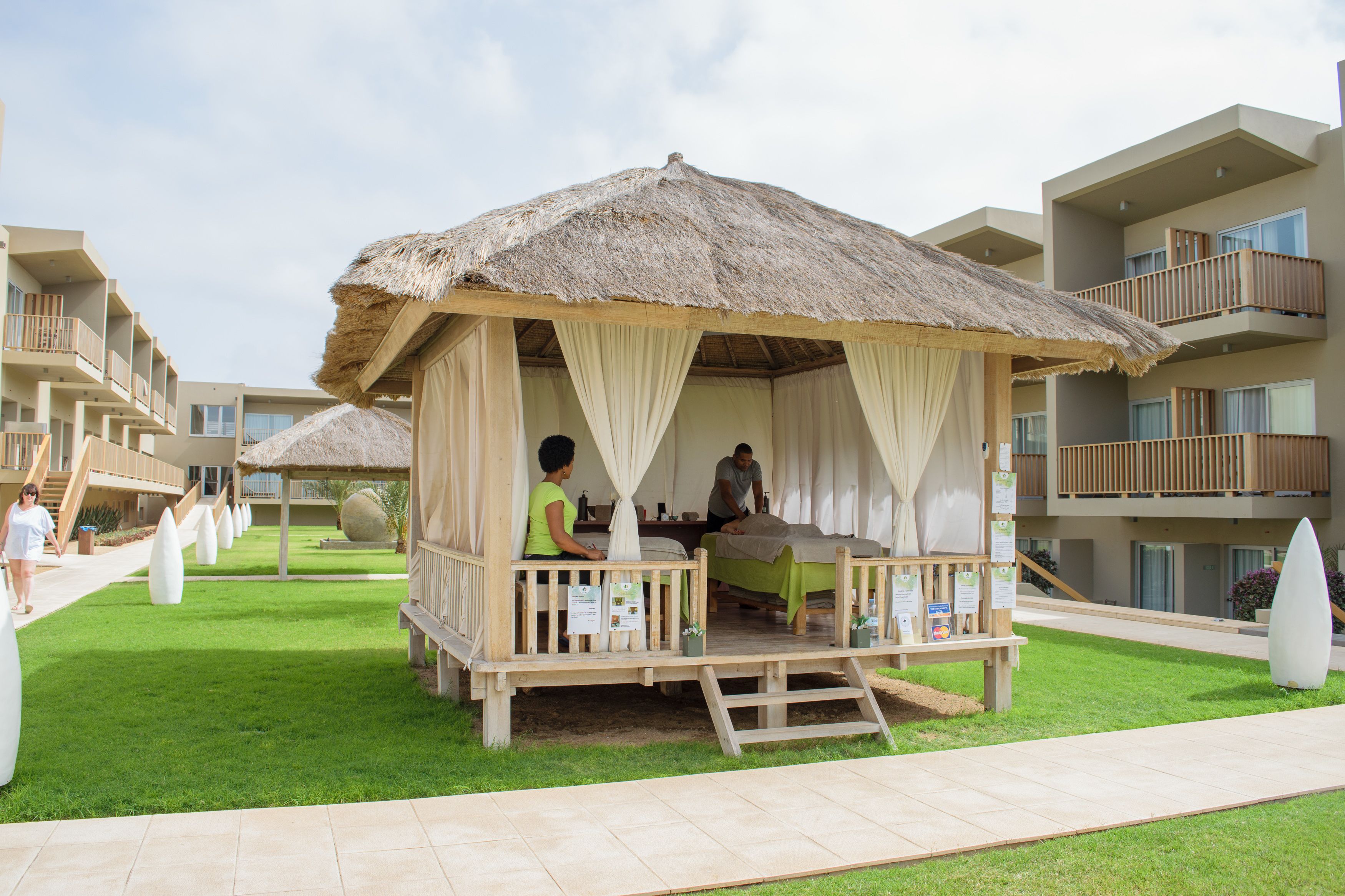 Un área de estar al aire libre con techo de paja, que incluye una banca de madera y una mesa, rodeada por un césped cuidado y un edificio moderno con balcones.