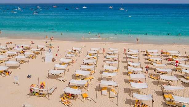 Een strand met veel ligbedden en parasols, helderblauwe zee en boten in de verte.
