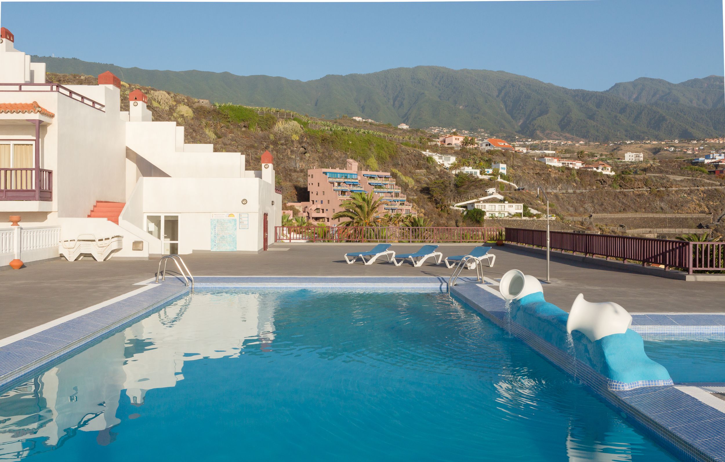 Uma piscina com água azul cristalina, rodeada por espreguiçadeiras e com vista para montanhas ao fundo.