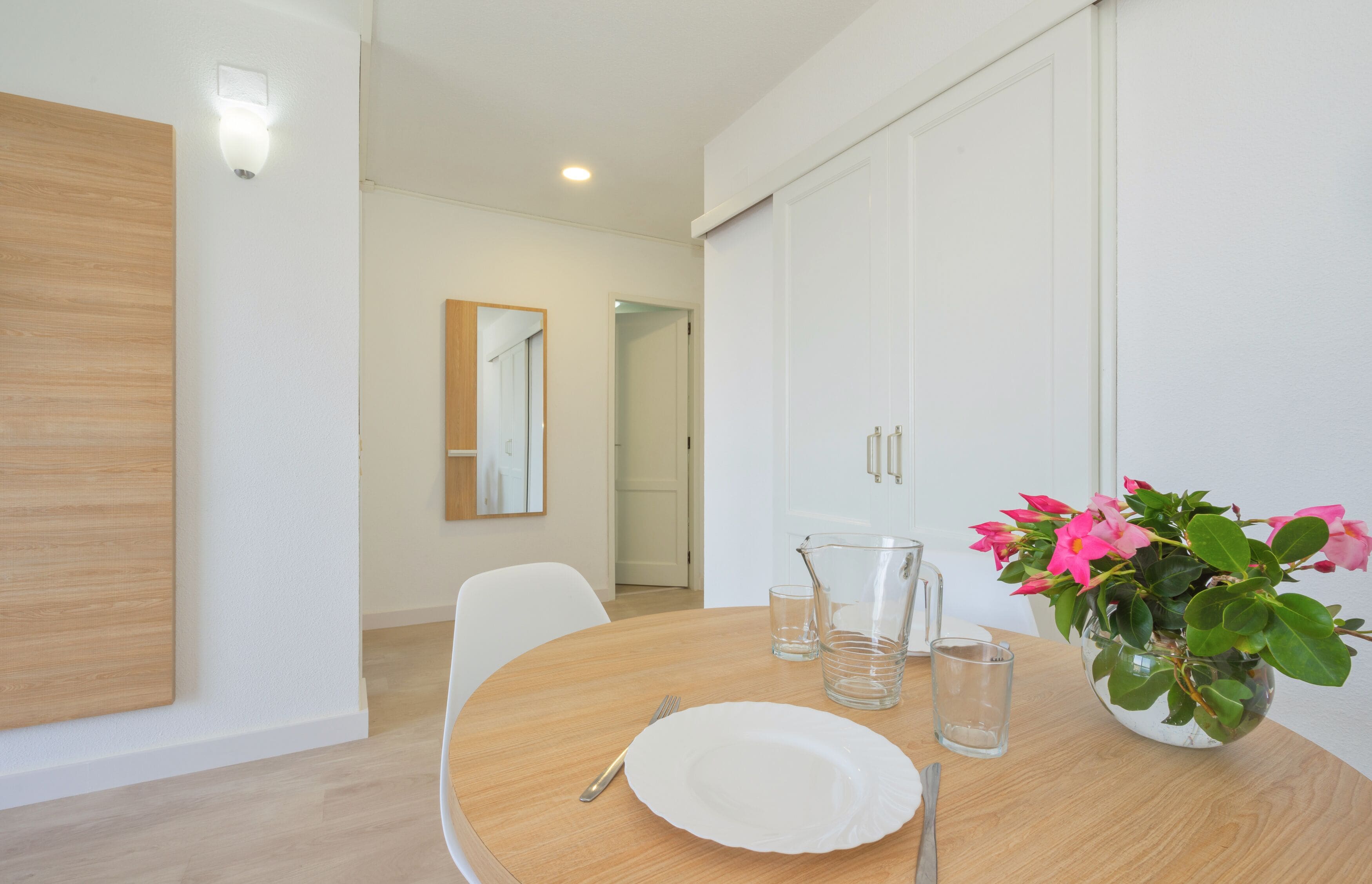 A dining area with a wooden table, white chairs, a white plate, a pitcher, and a vase with flowers.