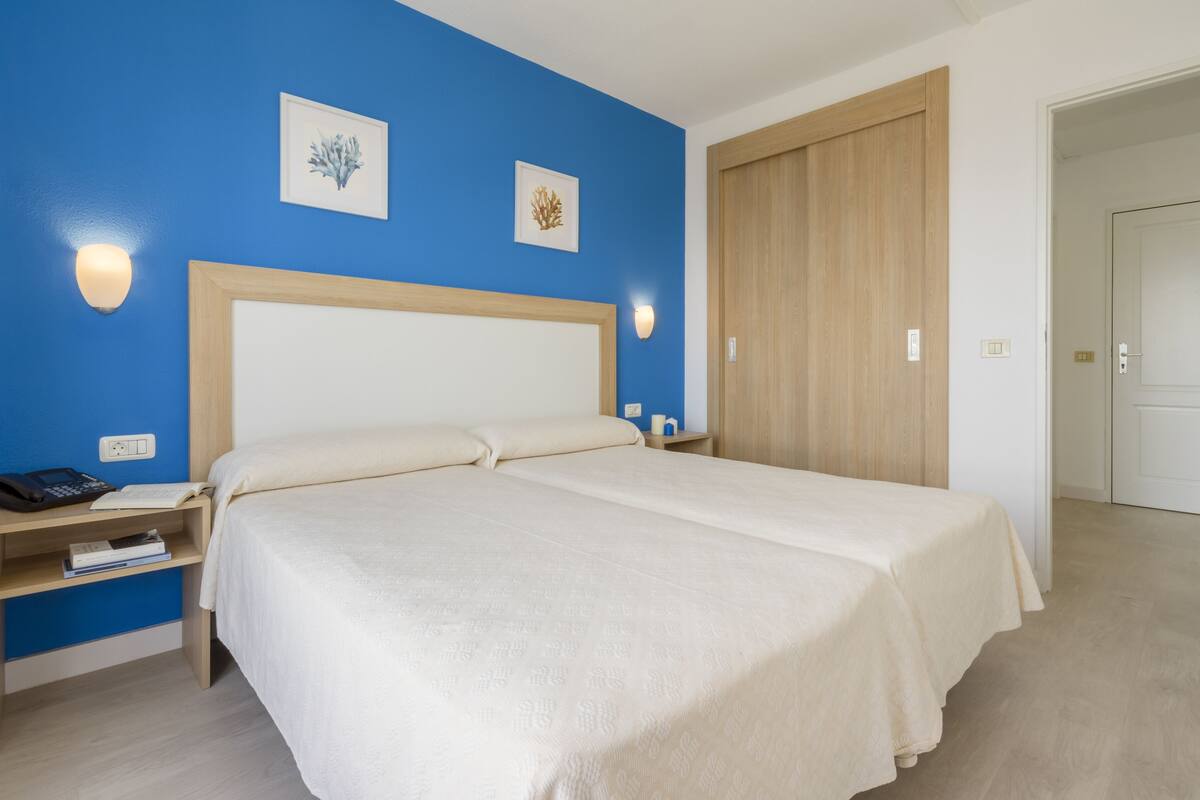 A hotel room with a large bed, a nightstand with a phone and books, and a wooden wardrobe.