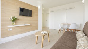 Una sala de estar moderna con una televisión de pantalla plana montada en una pared de madera, una mesita redonda con un libro, un sofá y un comedor con sillas blancas.