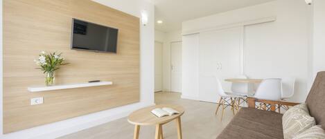 A modern living room with a flat-screen TV mounted on a wooden wall, a small round table with a book, a sofa, and a dining area with white chairs.