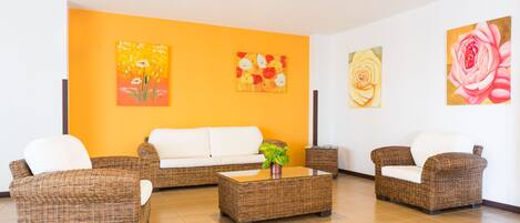 A living room with a white sofa, wicker chairs, a glass coffee table, and three floral paintings on the wall.