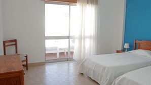 A hotel room with two beds, a wooden desk, a chair, a nightstand with a lamp, and a view of a balcony with a table and chairs. - Atlantic Side (Silves)