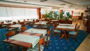 Un comedor de hotel con varias mesas y sillas, ventanas amplias y plantas en macetas.